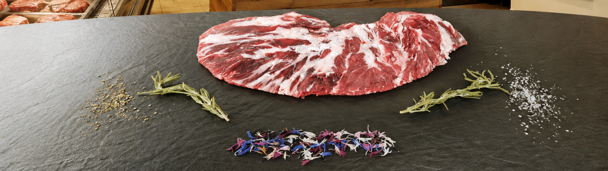 Rohes Rindersteak mit Marmorierung, daneben Rosmarinzweige, grobes Salz und bunte Blütenblätter auf schwarzem Stein